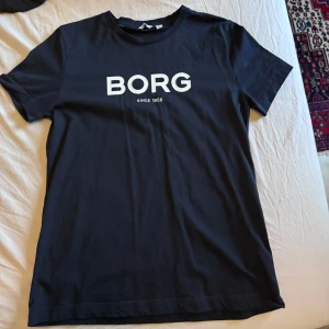 Blå t-shirt från Björn Borg - Snygg blå t-shirt från Björn Borg med vit text på bröstet. Klassisk rund halsringning och normal passform. Perfekt för en stilren och enkel look.