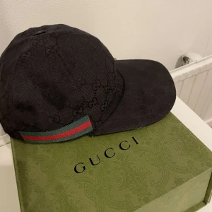 Gucci keps - Svart keps från Gucci med diskret GG-logomönster och klassiskt grönt och rött band på sidan. Kepsen är i storlek M (58 cm) och är i väldigt bra skick. Byten funkar och hör av er för fler frågor och den är äkta 
