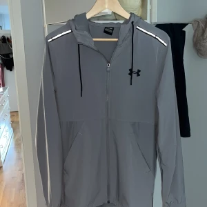 Grå träningsjacka från Under Armour - Säljer en grå träningsjacka från Under Armour med huva och dragkedja. Jackan har svarta snören, logga på bröstet och reflekterande detaljer på axlarna. Perfekt för träning och har en sportig look.