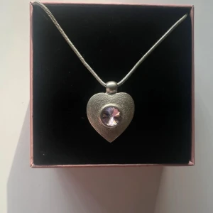 Halsband med hjärthänge och lila sten - Säljer ett silverfärgat halsband med ett hjärtformat hänge. Hänget har en glittrig lila sten i mitten som ger ett fint skimmer. Kedjan är tunn och justerbar med förlängningskedja. Perfekt accessoar för att lyfta din outfit. Asken medföljer ej, ca 44cm och helt oanvänt