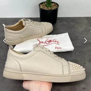 Beige sneakers med nitar från Christian Louboutin - Säljer ett par beige sneakers från Christian Louboutin med ikoniska röda sulor och coola nitar på tån. Skorna har snörning och är tillverkade i skinn. Perfekta för dig som vill sticka ut med en lyxig och edgy stil. Självfallet är dem äkta.