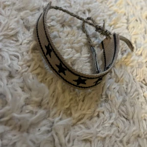 Beige tygarmband med svarta stjärnor - Säljer ett beige armband i tyg med svarta stjärnor som mönster. Armbandet har en flätad knytning och tofs i ena änden. Perfekt accessoar för att ge din stil en cool touch.