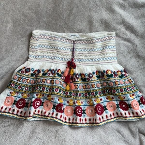 Broderad kjol med volang från Zara - Superfin vit kjol från Zara med broderade blommor i rött, rosa, gult och blått. Kjolen har volang och knytband med tofsar framtill. Perfekt för sommarens alla tillfällen och ger en bohemisk känsla. Använd 1-2 gånger. Nypris ca. 400:-