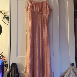 Ljusrosa långklänning med spetsdetaljer - Säljer en romantisk ljusrosa långklänning med smala axelband och vackra spetsdetaljer. Klänningen har en rak passform och är täckt av spetsmönster i vertikala ränder. Perfekt för somriga tillfällen eller studentfirande.