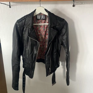 Svart skinnjacka från Jofama by Kenza - Säljer en svart skinnjacka från Jofama by Kenza i storlek 34. Jackan har klassisk bikerstil med sned dragkedja, bälte i midjan och quiltade detaljer på axlarna. Fodret är blommigt och färgglatt vilket ger en snygg kontrast.
