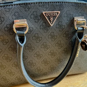Svart handväska från Guess - Elegant svart handväska från Guess med diskret logomönstet och lyxiga guldfärgade detaljer. Väskan har dubbla handtag, en avtagbar axelrem och flera smarta innerfack som håller dina saker organiserade. 