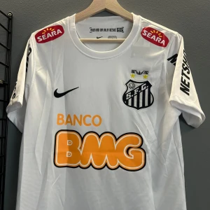 Vit Santos FC fotbollströja Neymar Jr - Vit kortärmad Santos FC matchtröja med svarta detaljer, sponsorloggor och Neymar Jr samt nummer 11 på ryggen. Klubbmärket är broderat på bröstet och tröjan har tryckta loggor på ärmar och bröst. En replika helt ny i storlek S