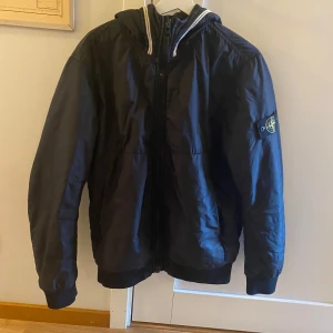 Svart dunjacka från Stone Island - Säljer en svart vindjacka från Stone Island med huva och dragkedja framtill. Jackan har en klassisk patch med logga på vänster ärm och sportiga detaljer vid huvan. Nypris 7000kr 