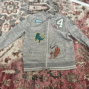 Grå ziptröja med djurpatches för små varn ca 8-10 - Grå ziptröja med dragkedja och coola patches, bland annat en tiger och ett lejon på framsidan. Tröjan har ribbade muddar och detaljer som siffran 4 och NYC-tryck. Perfekt för en avslappnad stil. Storlek 134-140 för små barn