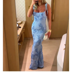 Ljusblå långklänning med blommigt mönster - Supersnygg ljusblå långklänning med blommigt mönster och smala axelband som knyts och går att justera på axlarna. Figurnära passform och fyrkantig urringning. Perfekt för sommarens alla tillfällen! Den är helt ny, aldrig använd! 