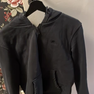 Svart hoodie med dragkedja från Burberry - Säljer en svart hoodie från Burberry i storlek M. Tröjan har dragkedja framtill, huva med snörning och klassiskt rutigt foder på insidan. Perfekt för dig som gillar stilrena märkesplagg med diskreta detaljer.