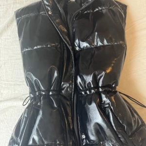 Glansig svart dunväst med dragkedja från H&M - Säljer en svart glansig dunväst med dragkedja och snörning i midjan. Västen har hög krage och är perfekt för att ge en cool look till din outfit. Passar lika bra till jeans som till kjol.