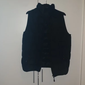 Svart dunväst från Zavetti Canada - Säljer en svart dunväst från Zavetti Canada med broderad logga på kragen och reflekterande detaljer. Västen har dragkedja framtill och justerbar dragsko nedtill. Perfekt för lager-på-lager under kyliga dagar.