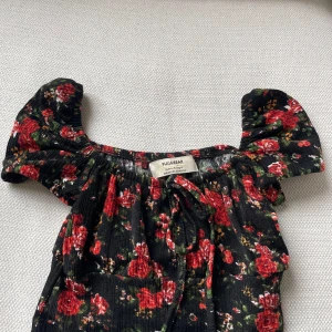Svart blommig croppad topp från Pull&Bear - Supersöt svart croppad topp från Pull&Bear med röda och gröna blommor. Toppen har korta puffärmar och en smockad, stretchig struktur. Perfekt för sommardagar och att styla med jeans eller kjol.