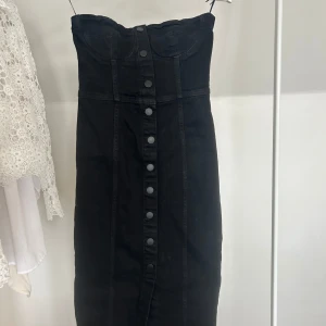Svart axelbandslös jeansklänning från Zara - Säljer en svart axelbandslös jeansklänning från Zara med markerad byst och knappar framtill. Klänningen har en figurnära passform och är tillverkad i ett kraftigare denimtyg. Perfekt för dig som gillar en stilren och edgy look.