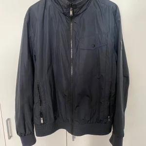  vindjacka från Hugo Boss - Säljer en mörkblå vindjacka från Hugo Boss med dragkedja framtill, två sidofickor med dragkedja och en bröstficka med knapp. Jackan har ribbade muddar och diskret BOSS-logga på bröstet. Perfekt för blåsiga dagar.