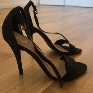 Svarta höga klackar med öppen tå - Säljer ett par svarta klackskor med öppen tå och smal rem runt vristen. Skorna har hög klack och enkel, stilren design som passar perfekt till festliga tillfällen. Spänne i metall vid ankeln för justerbar passform.