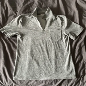 Ljusgrå pikétröja med dragkedja - Säljer en ljusgrå pikétröja med korta ärmar och dragkedja i halsen. Tröjan har en bröstficka och är i mjukt material, perfekt för en avslappnad stil.