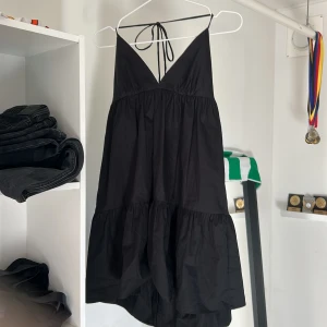 Svart kort klänning med volang och knyt i ryggen - Supersöt svart klänning med djup v-ringning och tunna axelband som knyts i nacken. Klänningen har volang nedtill och är perfekt för sommardagar. Lätt och luftig modell med öppen rygg.