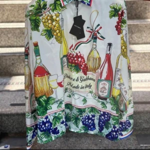 Färgglad skjorta med vinmotiv från Dolce & Gabbana - Säljer en unik skjorta från Dolce & Gabbana med färgglatt vinmotiv, druvor och flaskor. Skjortan har klassisk krage och knäppning framtill. Perfekt för dig som vill sticka ut med italiensk stil.