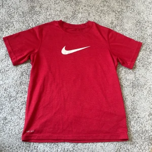 Röd Nike t-shirt med vit logga - Röd t-shirt från Nike med klassisk vit swoosh-logga på bröstet. Tillverkad i ett lätt och sportigt material med Dri-FIT-teknologi. Perfekt för träning eller en sportig vardagslook.