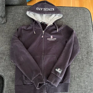 Marinblå hoodie från Kingsland Equestrian - Säljer en marinblå hoodie från Kingsland med dragkedja och huva. Tröjan har broderad logga på bröstet och tryck på huvan. Mjuk insida och praktiska fickor framtill. Perfekt för dig som gillar ridning och vill ha en snygg och bekväm hoodie.