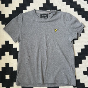 Grå t-shirt från Lyle & Scott - Säljer en stilren grå t-shirt från Lyle & Scott med klassisk rund hals och den ikoniska gula loggan broderad på bröstet. Perfekt basic-plagg som funkar till det mesta. Skön passform och mjukt material.