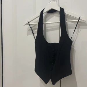 Svart väst med knappar - Snygg svart väst med djup v-ringning och knappar framtill. Västen har en figurnära passform och är croppad, perfekt att styla över en skjorta eller t-shirt.