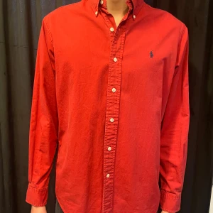 Röd skjorta från Ralph Lauren - Klassisk röd skjorta från Ralph Lauren med button-down krage och broderad logga på bröstet. Skjortan är tillverkad i 100% bomull och har långa ärmar samt vita knappar framtill.