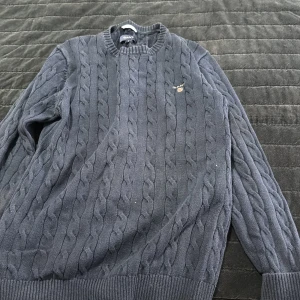 Mörkblå kabelstickad tröja från GANT - Säljer en klassisk mörkblå kabelstickad tröja från GANT. Tröjan har rund halsringning, ribbade muddar och ett diskret broderat GANT-logo på bröstet. Perfekt för lager-på-lager och snygg till jeans eller kjol.