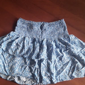 Blommig blå kjol från Kappahl - Charmig blå kjol från Kappahl med ett blommigt mönster. Kjolen har volanger som ger en lekfull känsla och en elastisk midja för bekväm passform. Perfekt för sommarens äventyr!