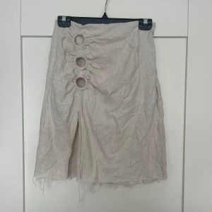 Beige kjol med ringdetaljer och slits - Unik beige kjol i linne med tre runda ringdetaljer framtill och en slits nedanför. Rå kant nedtill för en trendig look. Perfekt för dig som gillar en annorlunda och stilren stil.