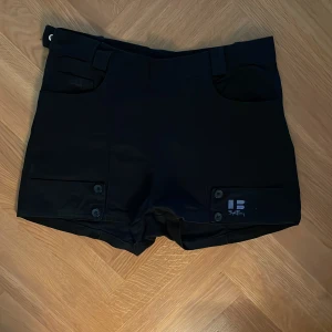 Svarta shorts från Björn Borg - Svarta shorts från Björn Borg. Modellen har hög midja, bälteshällor och två fickor framtill med knappdetaljer. 