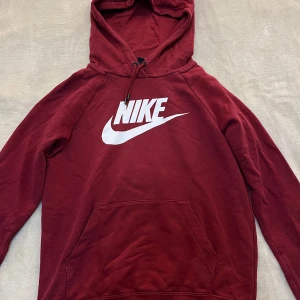 Vinröd hoodie från Nike - Säljer en vinröd hoodie från Nike med stor vit logga på bröstet. Tröjan har huva med dragsko och en stor magficka. Perfekt för dig som gillar sportig och avslappnad stil.