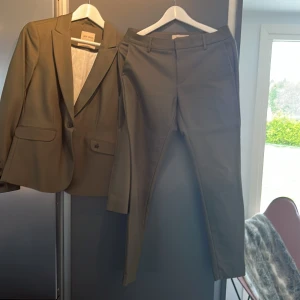 Olivgrön kostym från Mos Mosh - Snygg olivgrön kostym från Mos Mosh med klassisk kavaj och matchande byxor. Kavajen har en knapp och fickor med lock, byxorna har raka ben och bälteshällor. Perfekt för dig som vill ha en stilren och modern look.