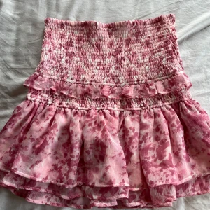Rosa volangkjol från Gina Tricot - Supersöt rosa kortkjol från Gina Tricot med volanger och smock i midjan. Kjolen har ett blommigt mönster och är perfekt för sommarens alla tillfällen. Lätt och luftig känsla med flera lager tyg. 
