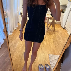 Mörkblå jeansklänning med smala axelband - Säljer en mörkblå, tight jeansklänning med smala axelband och markerade sömmar. Klänningen har en figurnära passform och är kort i modellen, perfekt för en trendig och avslappnad look. Passar dig som gillar denim och vill ha något enkelt men snyggt.