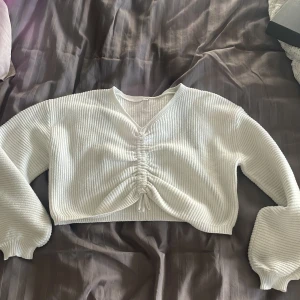 Vit croppad ribbad tröja med knyt - Säljer en vit croppad tröja med ribbad struktur och v-ringning. Tröjan har långa ärmar och snygg rynkdetalj med knyt framtill.