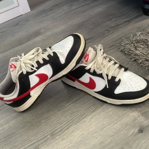 Nike Dunk Low med röd swoosh - Säljer ett par Nike Dunk Low sneakers i vitt och svart med röd swoosh och Nike-logga på plösen. Skorna har klassisk snörning och perforerad tå för extra ventilation. Perfekta för dig som gillar streetwear och snygga färgkombinationer.