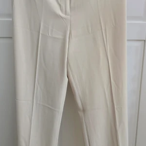 Beiga kostymbyxor med rak passform - Stilrena beiga kostymbyxor med rak passform och pressveck. Byxorna har hög midja och stängs med dragkedja och knapp. Perfekta för en clean och klassisk look.