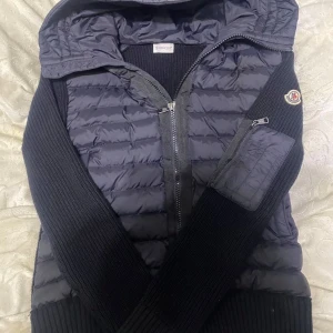  Moncler cardigan  - Snygg svart pufferjacka från Moncler med stickade ärmar och huva. Jackan har dragkedja framtill, en ficka med dragkedja på ärmen och Moncler-logga på axeln.       