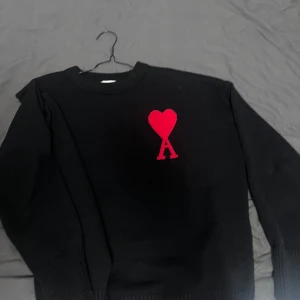 Svart stickad tröja med rött hjärta från Ami Paris - Svart stickad tröja från Ami Paris med ett rött hjärta och bokstaven A broderat på bröstet. Tröjan har rund halsringning och långa ärmar med breda ribbade muddar vid ärmslut och nederkant. Perfekt för en stilren och trendig look.