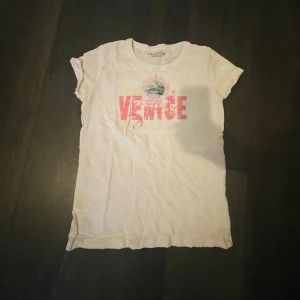 Vit t-shirt med rosa tryck  - Vit t-shirt med rosa tryck där det står 'Venice' och ett litet emblem ovanför. Klassisk rund halsringning och korta ärmar. Perfekt för en avslappnad stil.