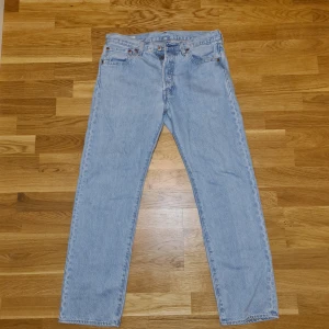 Levis 501 ljusblå jeans W32 L30 - Klassiska Levis 501 jeans i ljusblå denim med raka ben och knappgylf. Byxorna har fem fickor och den ikoniska Levis-lappen bak i midjan. Perfekta för en avslappnad och tidlös stil. W32 L30