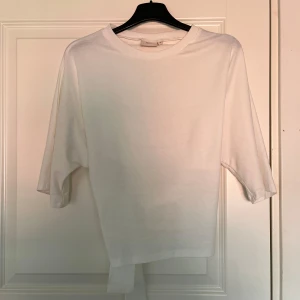 Vit topp med knyt i ryggen - Säljer en stilren vit topp med trekvartsärm och öppen rygg med knytband. Perfekt för dig som gillar enkla men snygga detaljer. Toppen har en rak passform och är gjord i mjukt bomullsmaterial.