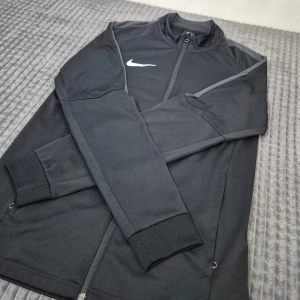 Svart träningsjacka från Nike - Säljer en svart träningsjacka från Nike med dragkedja och Dri-Fit-material. Jackan har långa ärmar, ribbade muddar och grå detaljer längs axlarna. Perfekt för dig som gillar sportig stil och vill ha något bekvämt till träningen.