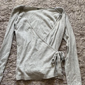 Ljusgrå omlottopp med knyt - Säljer en ljusgrå omlottopp med långa ärmar och knyt i sidan. Tunt ribbat material och djup v-ringning framtill. 💖