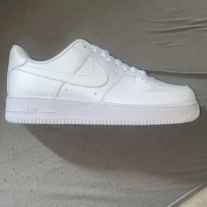Nike Air Force 1 helvita sneakers - Klassiska helvita Nike Air Force 1 sneakers med snörning och rund tå. Skorna har en platt sula och är tillverkade i skinn med diskreta detaljer och Nike-logga på hälen. Perfekta för en clean och stilren look.
