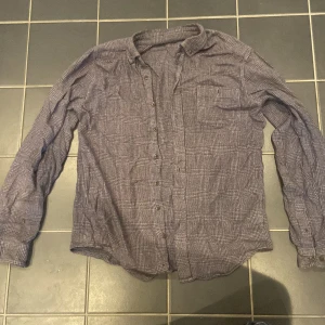 Rutig skjorta i grått och vitt - Säljer en klassisk rutig skjorta i grått och vitt med långa ärmar och knappar framtill. Skjortan har en bröstficka och är i mjukt tyg, perfekt för en avslappnad stil.