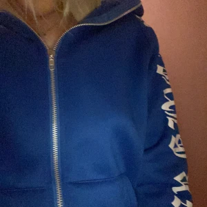 Blå hoodie med tryck på ärmarna - Säljer en blå hoodie med dragkedja och stora vita tryck längs båda ärmarna. Jackan har huva och två fickor framtill. Perfekt för en avslappnad och cool stil.